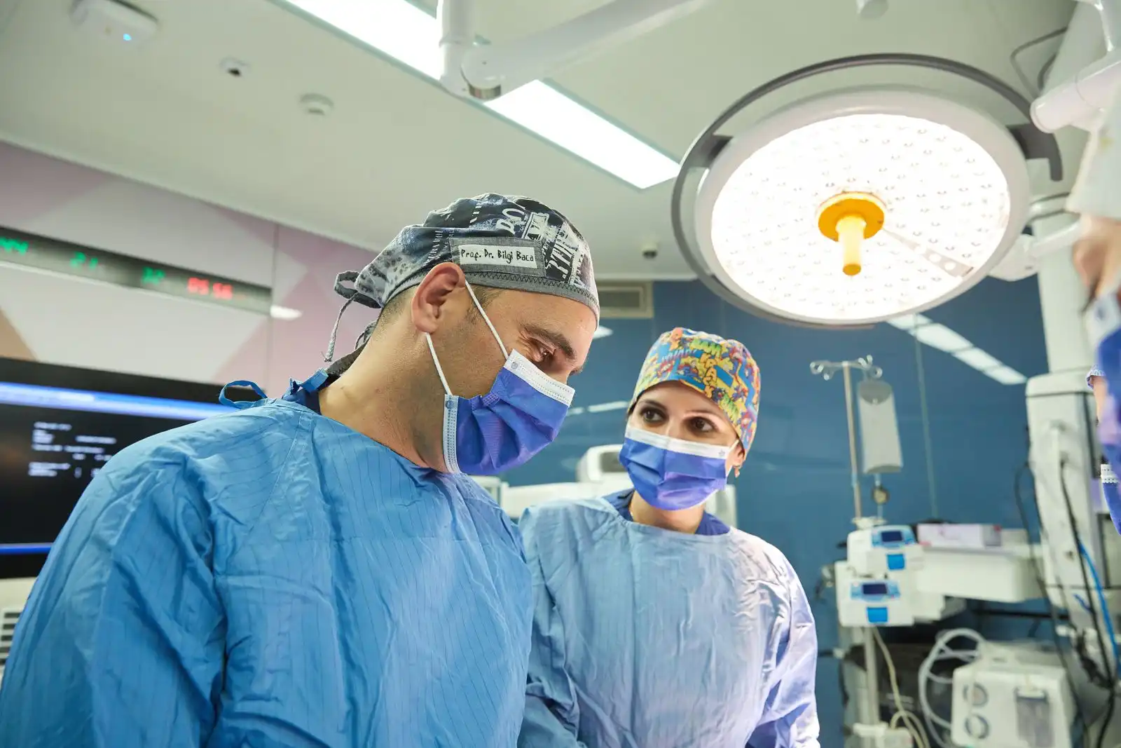 Prof. Dr. Bilgi Baca and surgical team member in blue scrubs and surgical masks performing plastic surgery under bright operating room lights, fully equipped modern operating theater in Turkey